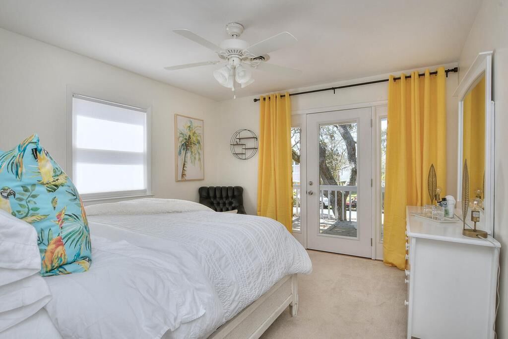 A bedroom with a bed, a ceiling fan, a window with blinds, a door with yellow curtains, and a dresser with toiletries.