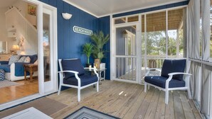 Eine überdachte Veranda mit zwei weißen Stühlen, eine blaue Wand mit einem Schild 'BEACH HOUSE' und Blick ins Freie.