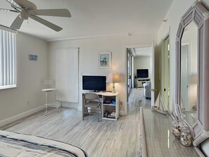 Habitación de hotel con ventilador de techo, un escritorio con computadora y lámpara, televisión, una silla y un espejo grande.
