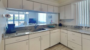 A modern kitchen with white cabinets, a marble countertop, a built-in dishwasher, and a large window with blinds. - Ocean Harbor by Distinctive Beach Rentals (Fort Myers Beach)