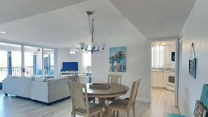 A modern living room with a dining area, a large window, and a kitchen in the background. - Ocean Harbor by Distinctive Beach Rentals (Fort Myers Beach)