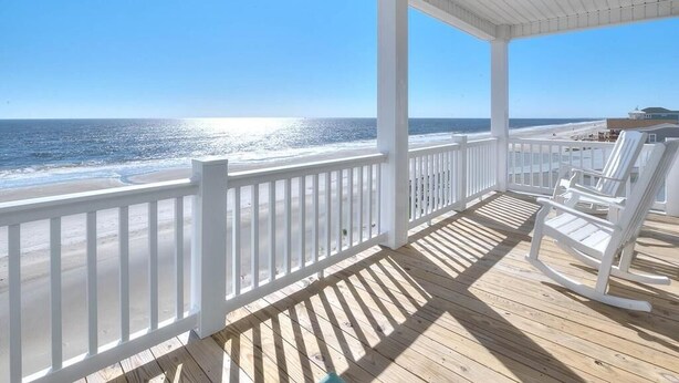 A wooden deck with white railings overlooking a beach. - Oceanfront Dory: Spacious 5BR, 4BA home in Oak Island, perfect for 10 guests. (Oak Island)