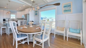 A spacious kitchen with white cabinetry, a wooden dining table surrounded by white chairs, and a view of the ocean through large windows. - Oceanfront Dory: Spacious 5BR, 4BA home in Oak Island, perfect for 10 guests. (Oak Island)