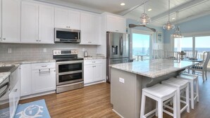 A kitchen with white cabinets, stainless steel appliances, a large island with stools, and a view to the outside. - Oceanfront Dory: Spacious 5BR, 4BA home in Oak Island, perfect for 10 guests. (Oak Island)