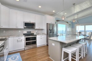 A kitchen with white cabinets, stainless steel appliances, a large island with stools, and a view to the outside.