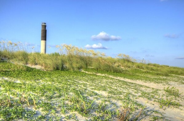 Paisagem costeira com um farol, dunas de areia e vegetação rasteira.