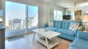 Un salon moderne avec un canapé bleu, une table basse en bois et une grande porte-fenêtre coulissante donnant sur un balcon aménagé avec du mobilier d’extérieur.