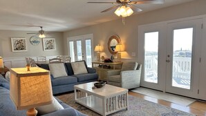 A living room with a blue sofa, a white coffee table, and a ceiling fan. There are two doors leading to a balcony with a view.