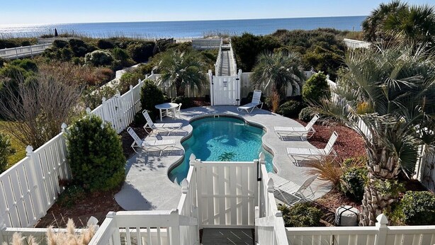A pool with a wooden deck, surrounded by white fences and palm trees, overlooking a beach.