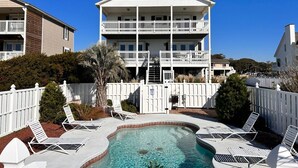 A two-story building with a balcony, a swimming pool with lounge chairs, and a white fence.