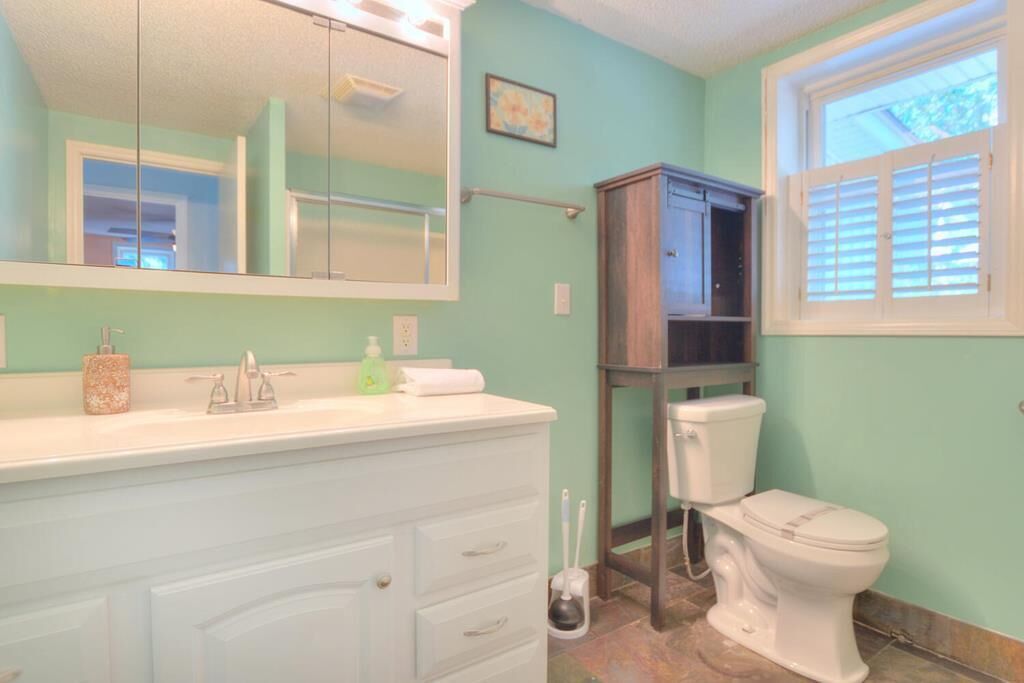 Une salle de bain avec un meuble-lavabo blanc, un miroir, des toilettes et une fenêtre avec des stores.