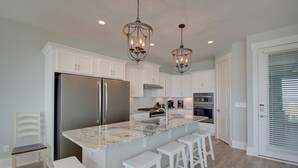 A modern kitchen with white cabinets, a stainless steel refrigerator, a marble island with bar stools, and pendant lighting. - Experience oceanfront luxury at AfterDune Delight, Oak Island's ideal getaway for 20 guests. (Oak Island)