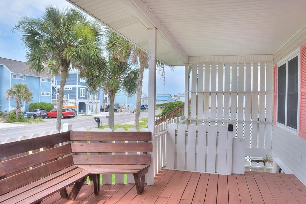 A wooden bench on a covered porch with a white fence and palm trees in the background.