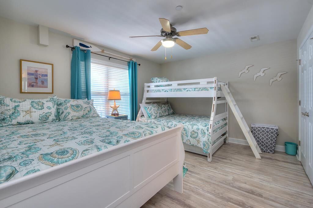 A bedroom with a bunk bed, a single bed, a ceiling fan, a window with curtains, a framed picture on the wall, and a trash bin.