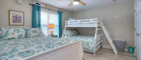 A bedroom with a bunk bed, a single bed, a ceiling fan, a window with curtains, a framed picture on the wall, and a trash bin.