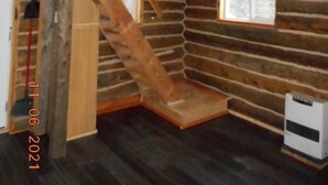 L’intérieur d’un chalet rustique, avec un escalier en bois menant à une mezzanine, une petite fenêtre et un radiateur posé sur le sol.