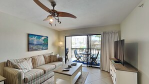 A living room with a sofa, coffee table, and a TV on a cabinet. There is a painting on the wall and a balcony with chairs and a table. - Santa Maria by Distinctive Beach Rentals (Fort Myers Beach)