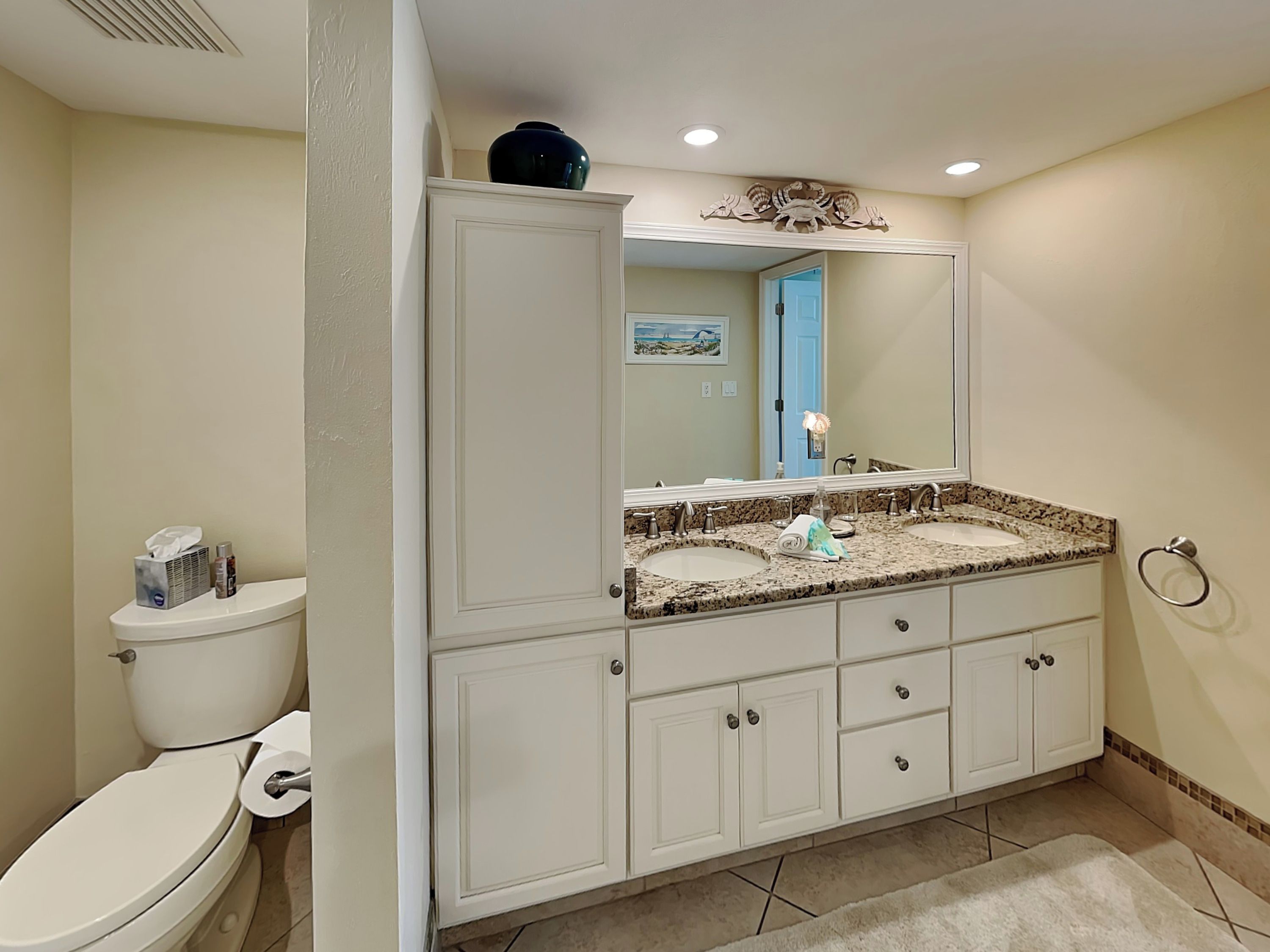 A bathroom with a white toilet, a double sink vanity with a large mirror, and a decorative wall piece above the mirror.