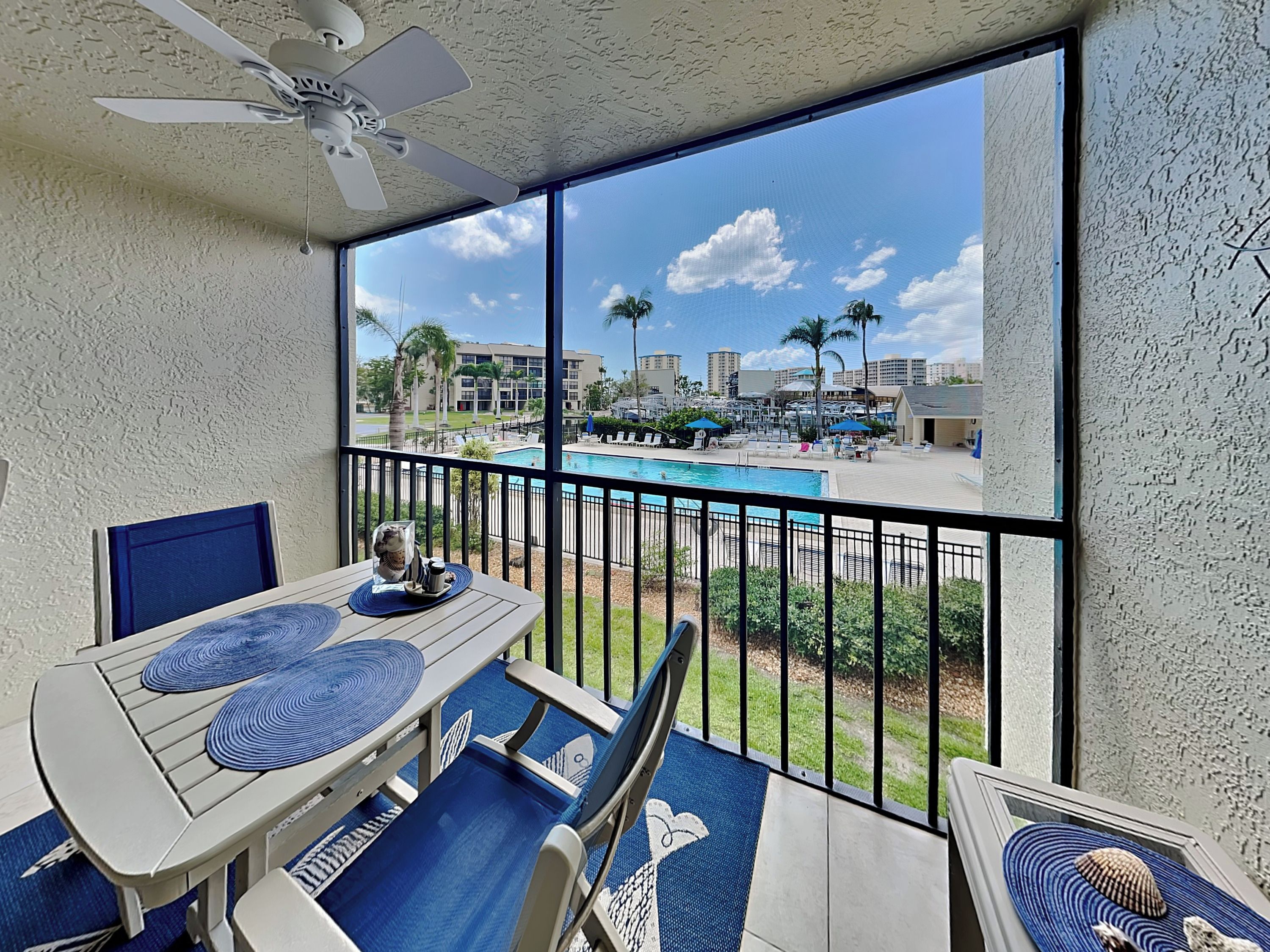 A balcony with a table and chairs, a ceiling fan, and a view of a swimming pool and buildings.