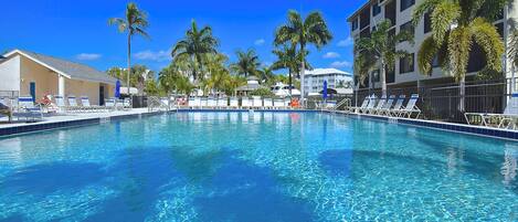A clear blue pool with lounge chairs and palm trees, surrounded by buildings.