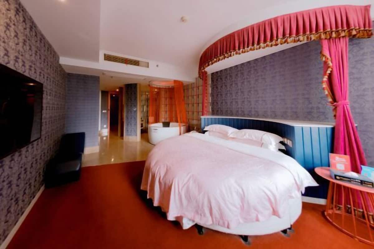 A hotel room with a large bed, a TV, a desk with a chair, and a bathroom visible in the background.