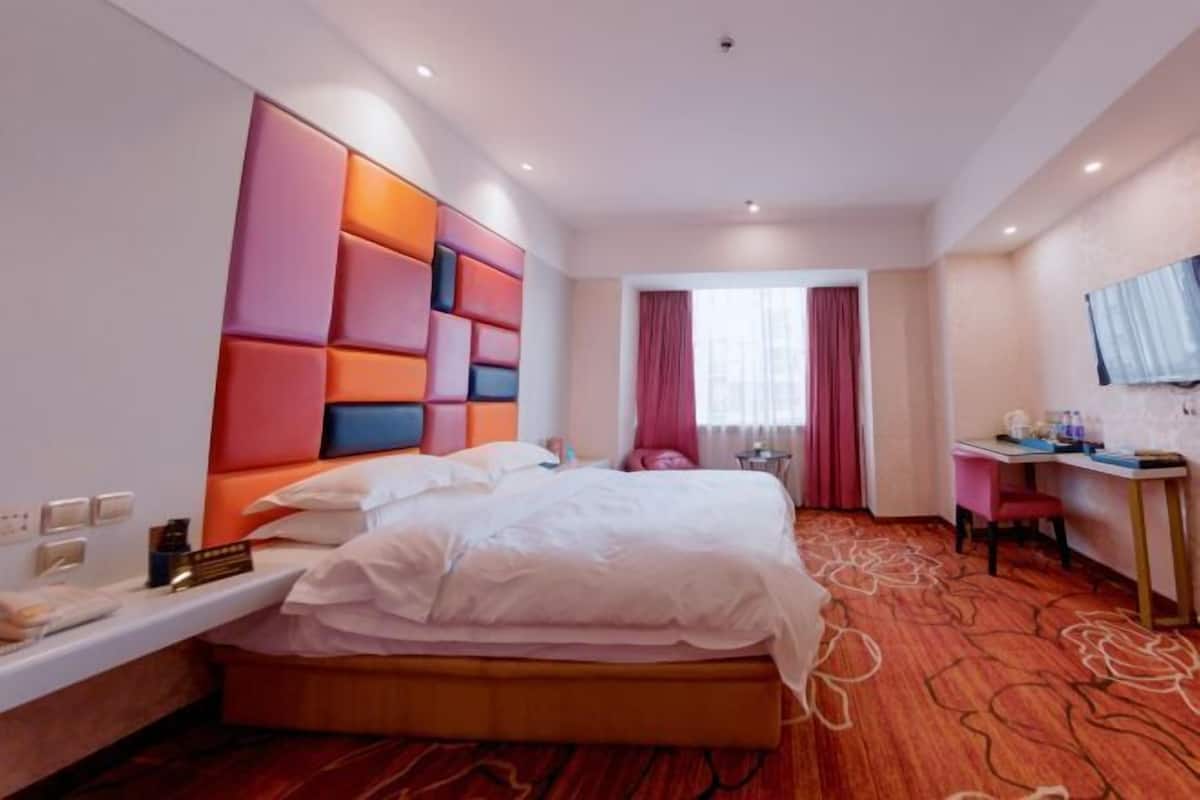 A hotel room with a bed, a desk, a chair, a window with curtains, and a wall-mounted TV.
