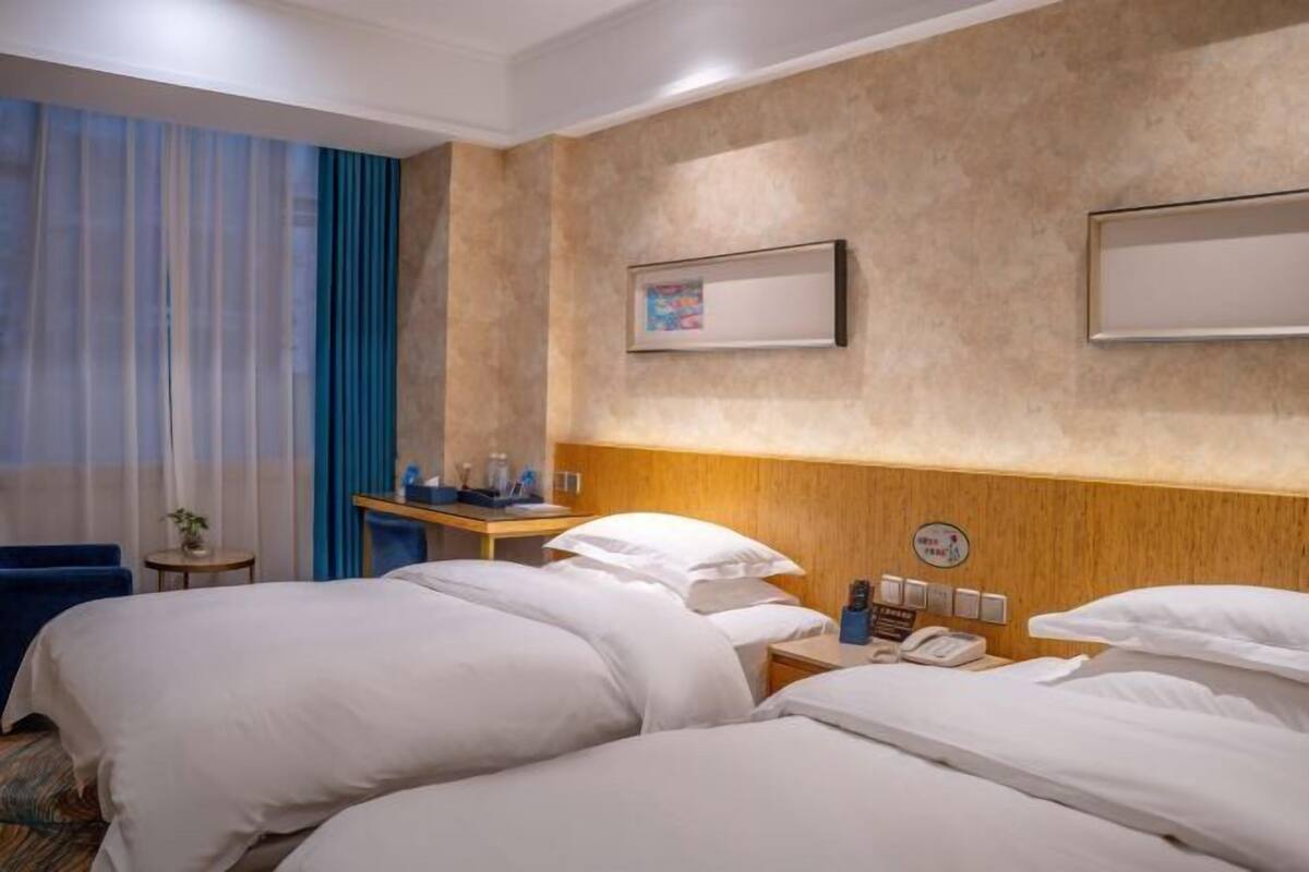 A hotel room with two beds, a bedside table with a phone, a small table with a plant, and a wall-mounted picture.