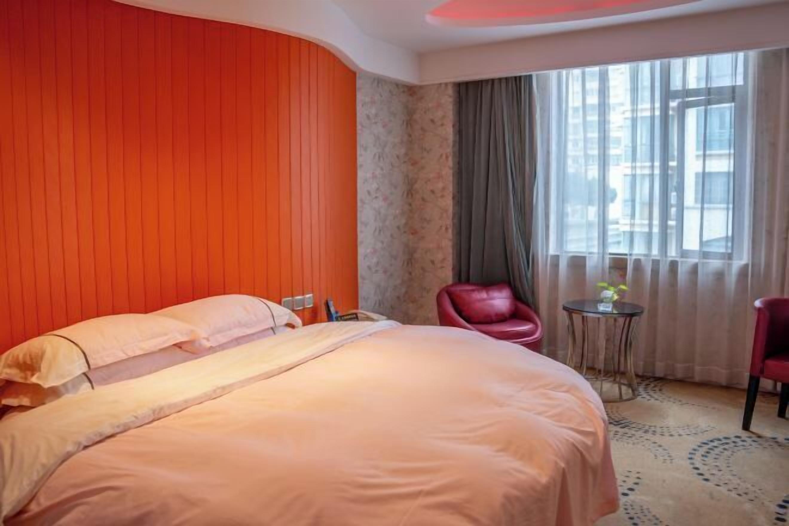 A hotel room with a large bed, a red chair, a small table, and a window with curtains.