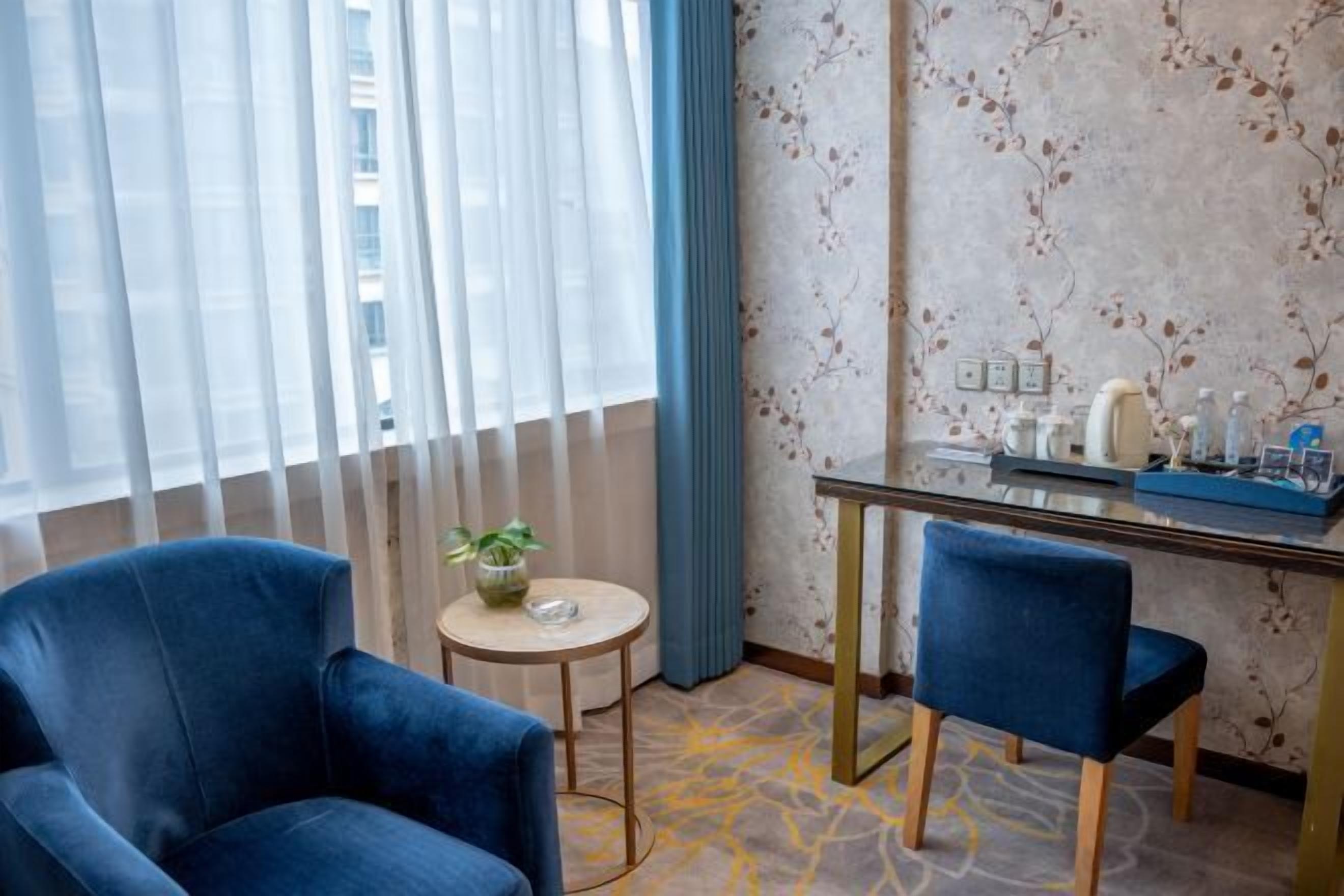 Habitación de hotel con dos sillones azules, una mesita redonda con un jarrón de flores, un escritorio con artículos de aseo y un papel tapiz estampado.
