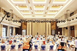Un amplio salón de conferencias con filas de mesas, un escenario con equipo de iluminación y un techo alto con un diseño de cuadrícula.