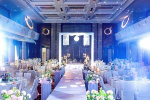 Un salón de banquetes con una larga mesa dispuesta para un evento formal, flanqueado por filas de sillas y mesas, con un pasillo central que conduce a un escenario.