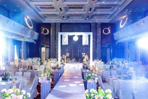 Un salón de banquetes con una larga mesa dispuesta para un evento formal, flanqueado por filas de sillas y mesas, con un pasillo central que conduce a un escenario.