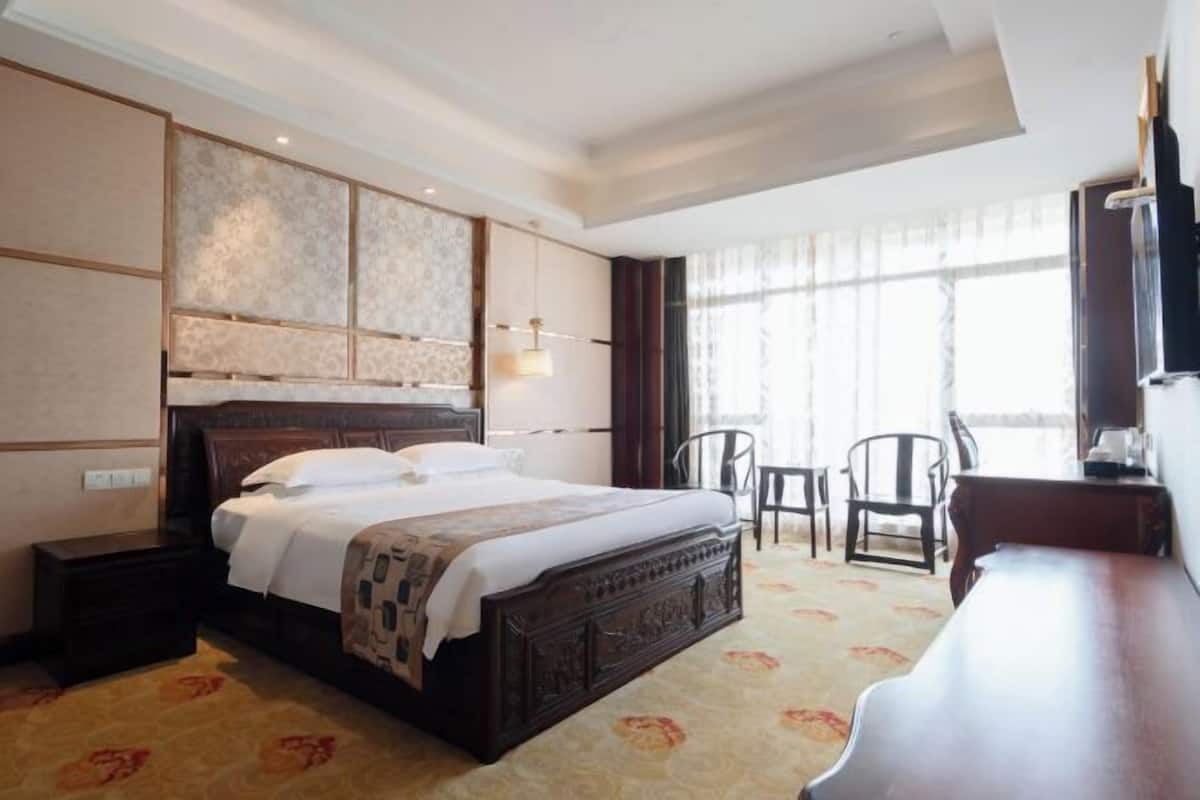 A hotel room with a large bed, a desk, two chairs, a TV, and a window with curtains.