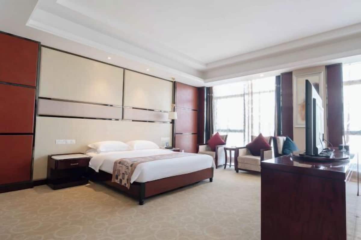 A hotel room with a large bed, a desk, and a chair. There is a window providing natural light and a view of the outside.