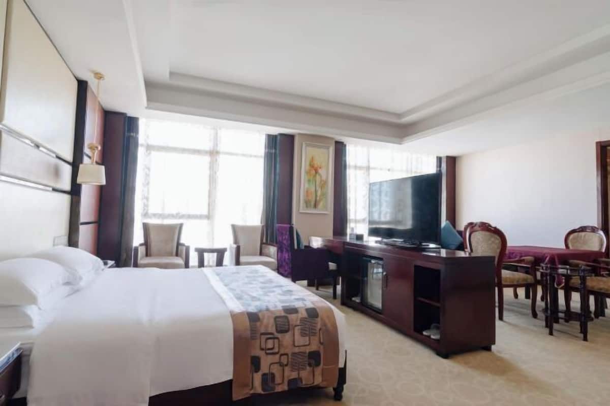 A hotel room with a bed, a desk with a television, a dining area with chairs, and a window providing natural light.