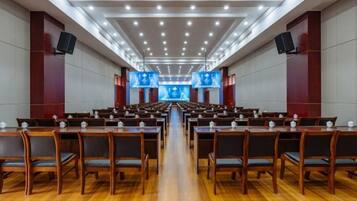 A conference room with rows of tables and chairs, large screens at the front, and a high ceiling with recessed lighting.