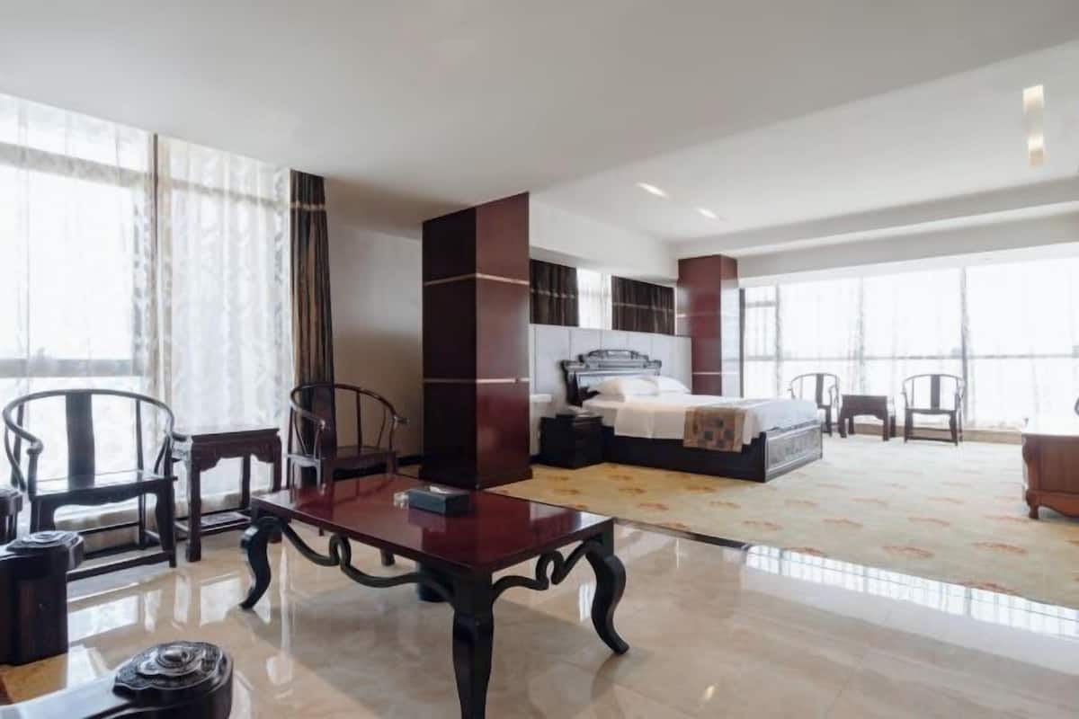 A spacious hotel room with a large bed, a sitting area with a table, and a view through large windows.