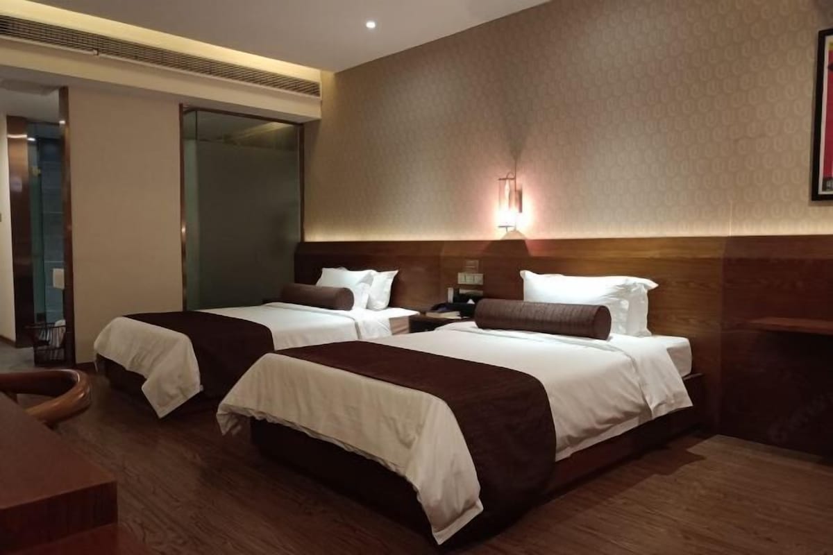 A hotel room with two beds, a wooden headboard, a bedside table, and a wall-mounted light fixture.