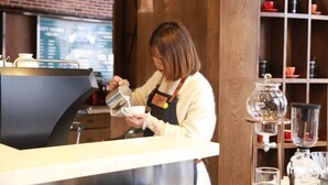 A barista at a counter with a coffee machine, a menu board, and shelves with cups and glasses.