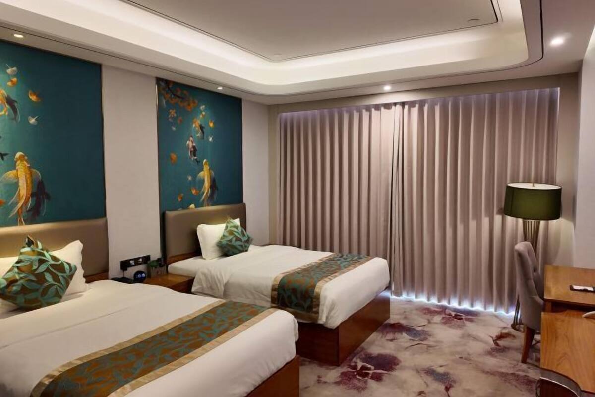 A hotel room with two beds, a large window with curtains, a desk with a lamp, and a painting on the wall.