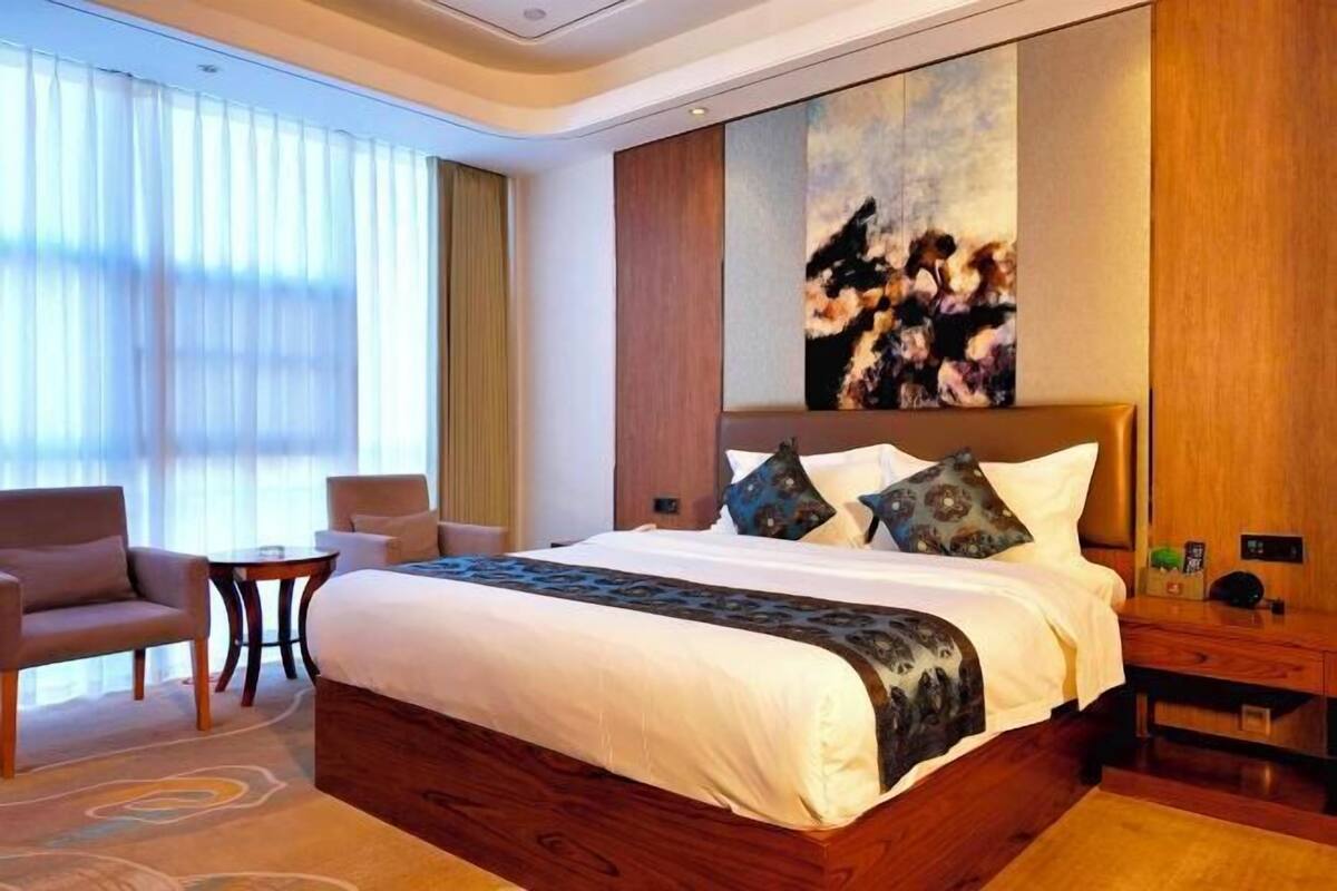 A hotel room with a large bed, a side table, a chair, and a painting on the wall.