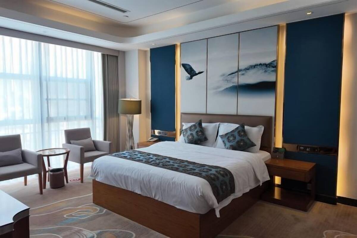 A modern hotel room with a large bed, a seating area with two chairs, a small table, and a large window with curtains.