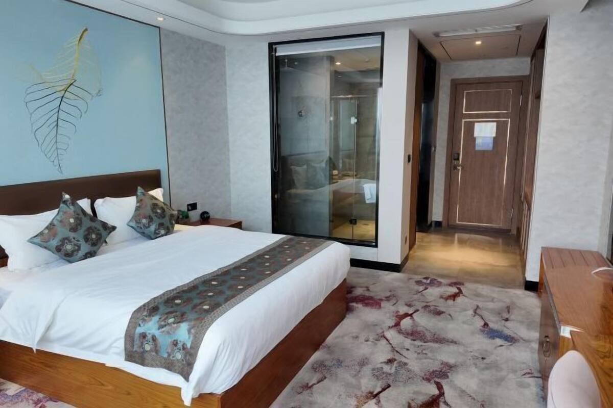 A modern hotel room with a large bed, a glass shower enclosure, a wooden door, and a view of the hallway.