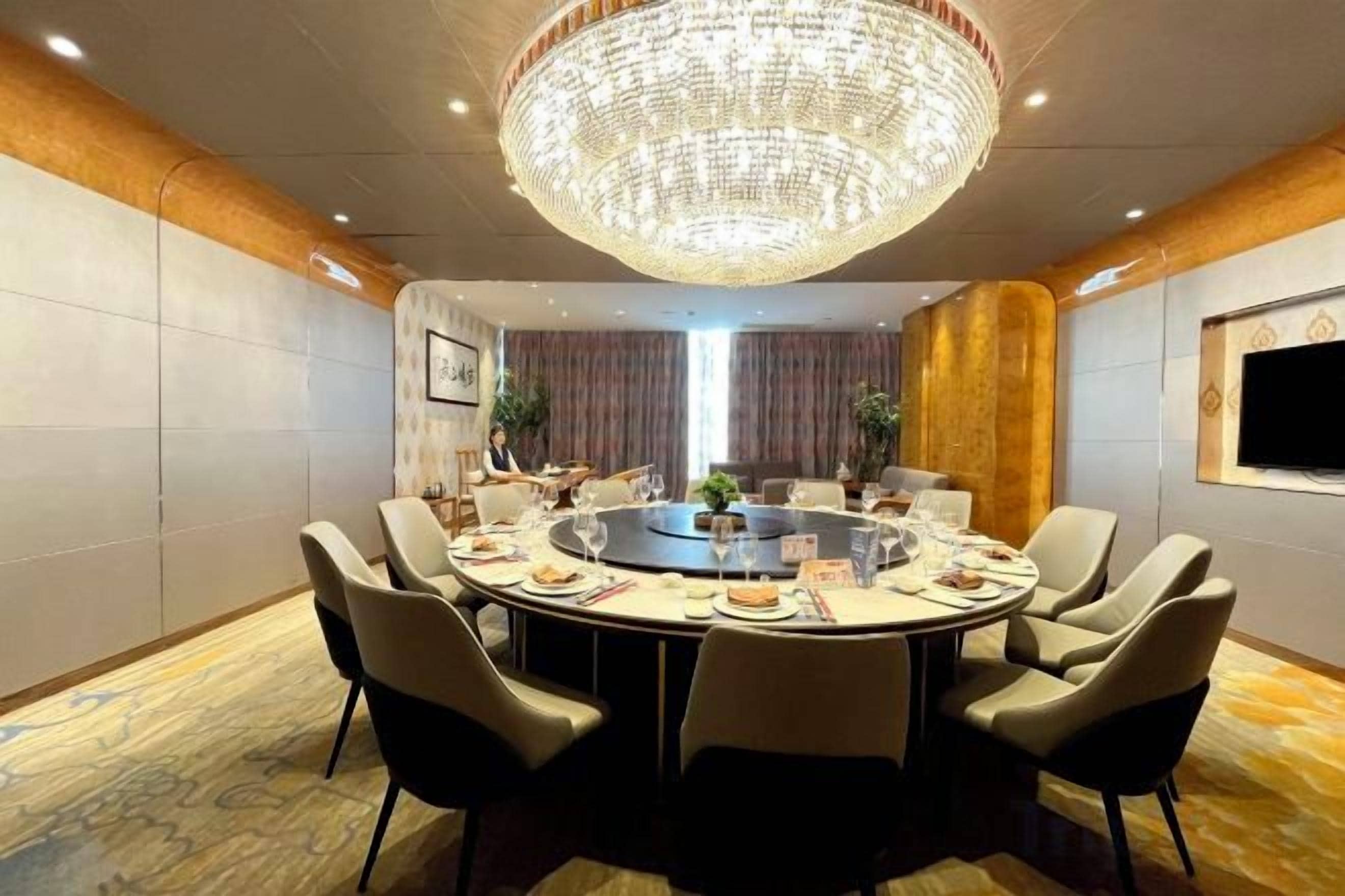 A conference room with a round table, chairs, a chandelier, and a television on the wall.