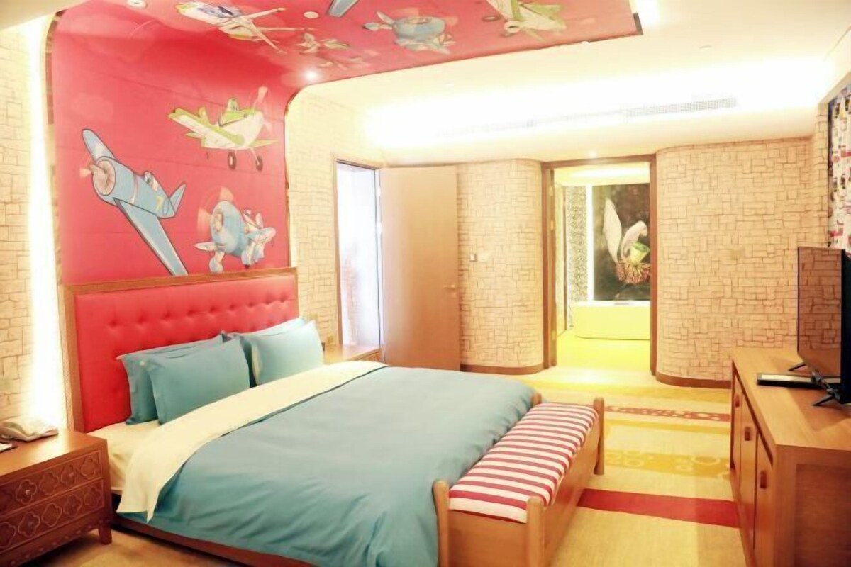 Un dormitorio con cabecera roja, cama con ropa de cama azul, un banco de madera, un televisor y una pared con diseño de ladrillo.