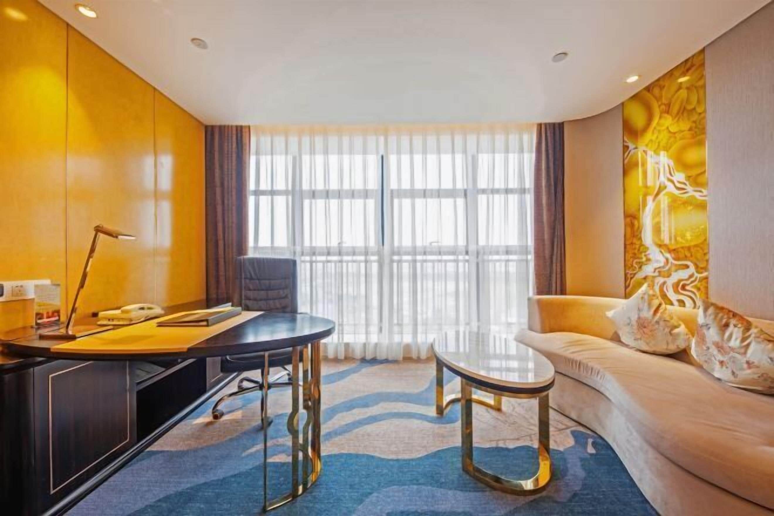 A modern hotel room with a desk, a round table, a sofa, and a large window with curtains.