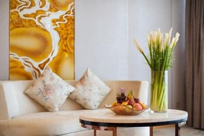 Una habitación con un sofá, una mesa redonda con frutero y un jarrón de flores, y un cuadro abstracto grande en la pared.
