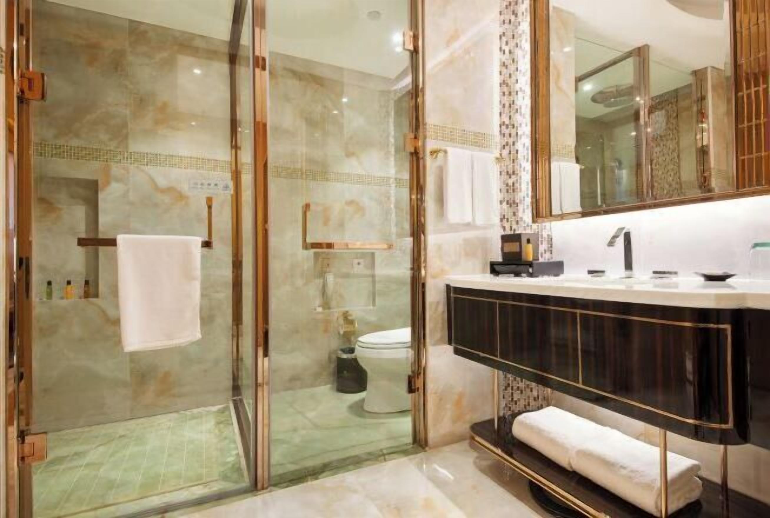 A modern bathroom with a glass-enclosed shower, a marble-tiled floor, a black vanity with gold accents, and a large mirror.
