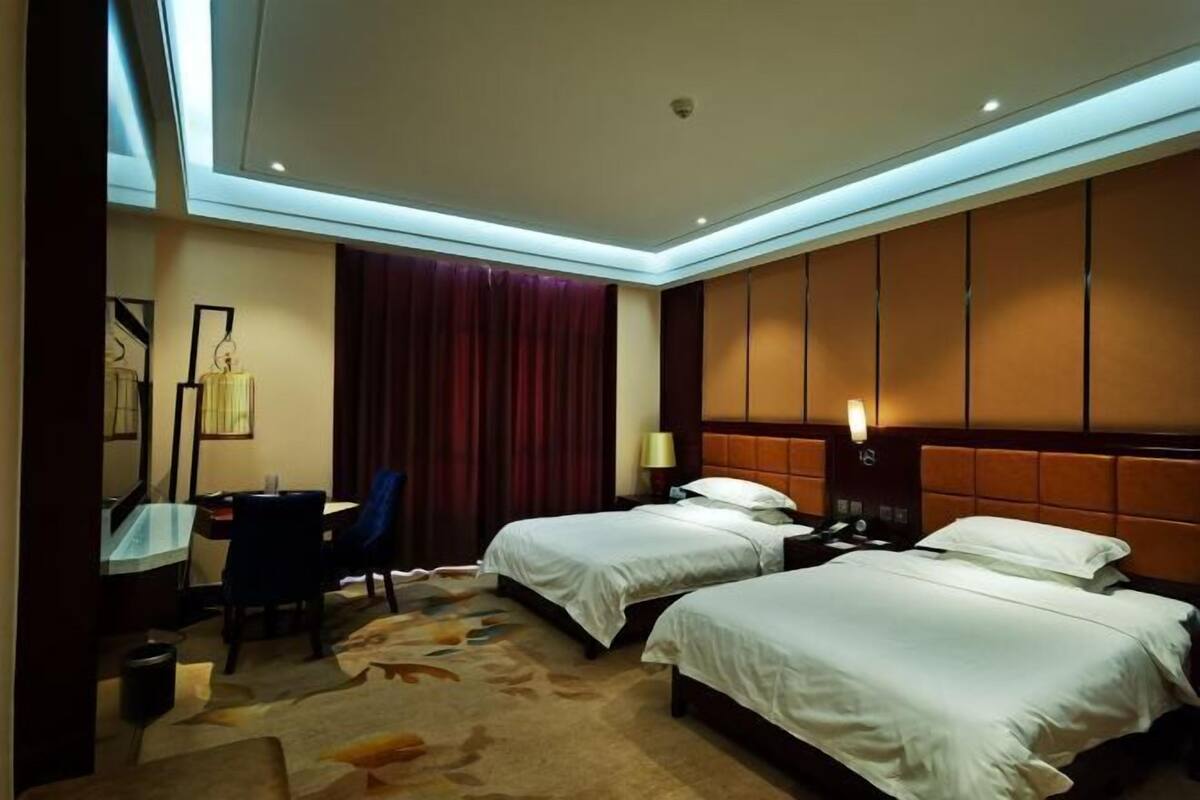 A hotel room with two beds, a desk with a chair, a desk lamp, and a large window with curtains.