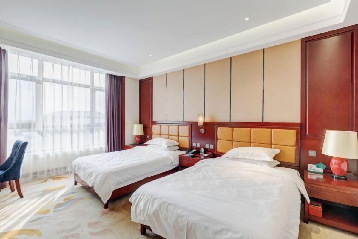 Habitación de hotel con dos camas, un ventanal con cortinas, una silla y una mesita de noche con lámpara.